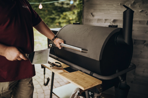 Mann bedient großen dunklen Grill im Garten mit Grillwerkzeugen auf Holztisch