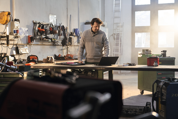 Mann in Werkstatt arbeitet konzentriert am Laptop umgeben von diversen Werkzeugen