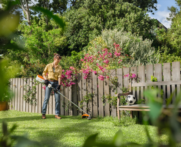Frau mittleren Alters mäht Rasen mit Stihl FSA 80 R Motorsense in einem blühenden Garten
