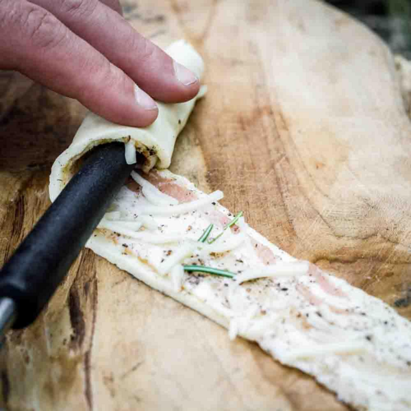 Hand wickelt Teig mit Schinken und Kräutern um Gusseisenspitze eines Stockbrot Spießes auf Holzbrett