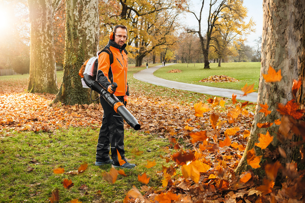 Mann in orangener Jacke benutzt STIHL Laubbläser in einem herbstlichen Park