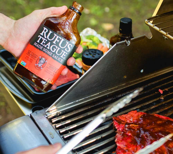 Hand hält Rufus Teague Blazin Hot BBQ Sauce über gegrilltem Fleisch