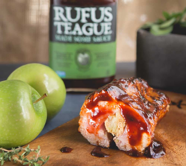 Schweinefleisch mit Rufus Teague BBQ-Sauce auf Holzbrett neben Äpfeln und Sauceflasche