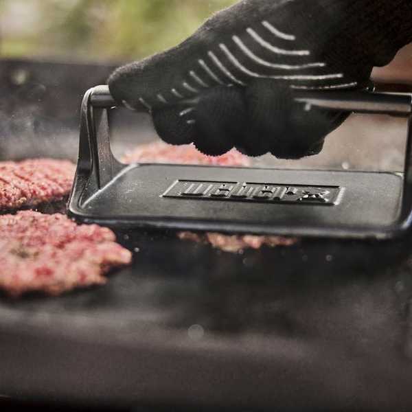Hand in hitzebeständigem Handschuh verwendet Weber Hamburgerpresse auf Grillplatte