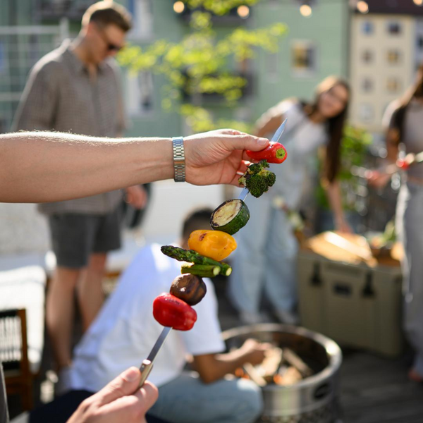 Person hält Gemüse-Grillspieß bei sonniger Grillparty auf Terrasse