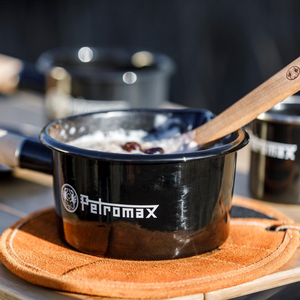 Schwarzer Petromax Emaille Stieltopf mit Essen und Holzlöffel auf orangefarbenem Untersetzer