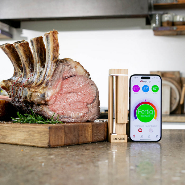 Smartes Kochthermometer MEATER Pro neben perfekt gegartem Rippenstück und Smartphone mit App-Anzeige
