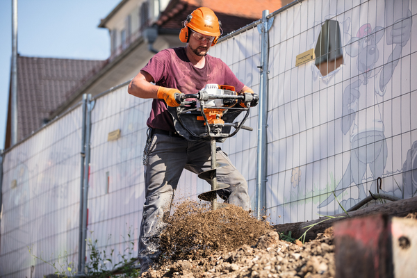 Mann verwendet STIHL BT 131 Erdbohrgerät auf Baustelle mit Sicherheitsausrüstung und Cartoon-Plane im Hintergrund
