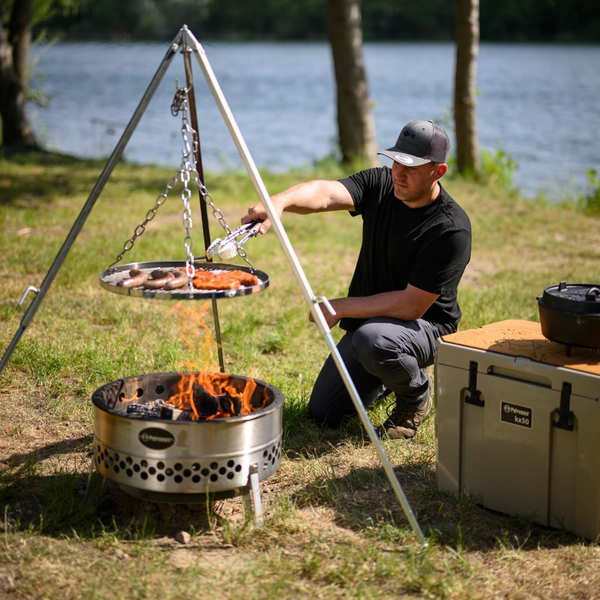 Mann grillt Fleisch über Petromax Feuerschale am Seeufer