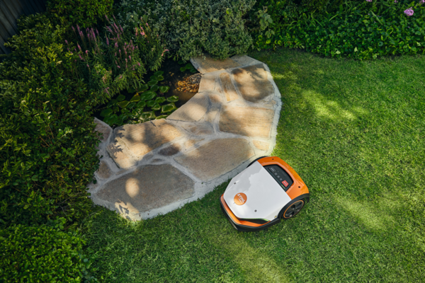 STIHL iMOW 7 Mähroboter mäht selbstständig einen grünen Rasen umgeben von Blumen und einem Teich