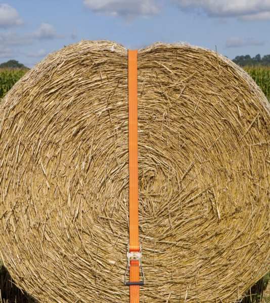 Orangefarbener Ratschenzurrgurt sichert großen Strohballen auf Feld