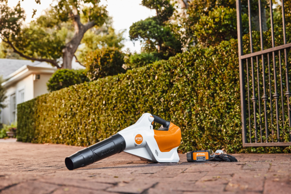 STIHL BGA 160 Akku-Laubbläser auf einem Weg vor einem Haus mit Hecke und Handschuhen