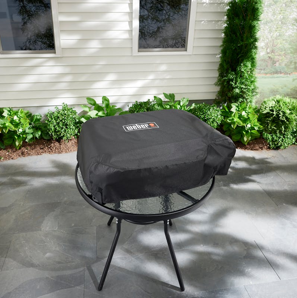 Schwarze Weber Premium Abdeckhaube auf runder Plancha auf Terrasse mit grünen Pflanzen