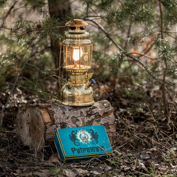 Petromax HK500 Petroleumlampe in einem Wald auf einem Baumstumpf neben einer blauen Logo-Box