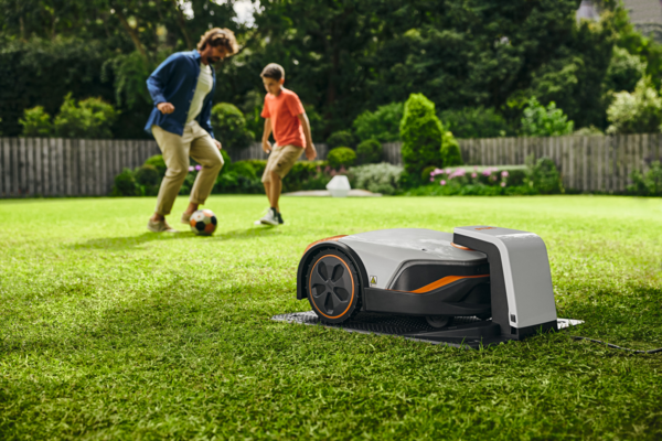 STIHL iMOW 6 Mähroboter im Einsatz auf Rasen mit spielendem Erwachsenen und Kind im Hintergrund