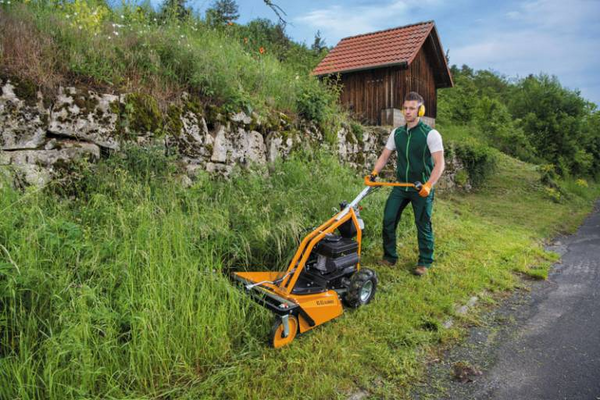Mann bedient AS 63 2T ES Rasenmäher auf steilem Grashang neben Steinwall und Holzhütte