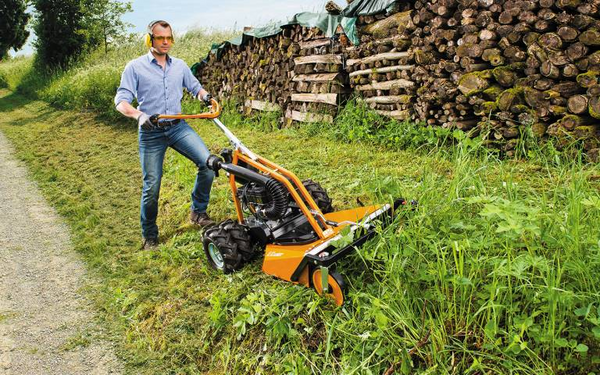 Mann bedient AS 63 2T ES Rasenmäher auf geneigter Wiese neben Waldweg