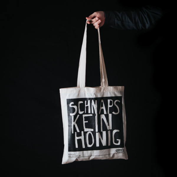 Person hält Jutebeutel mit Aufschrift 'Schnaps, kein Honig' vor schwarzem Hintergrund