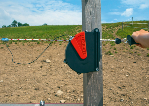 Flexigate Seilanschlusskabel an Holzpfosten auf Pferdekoppel mit bedienender Hand und Landschaft im Hintergrund