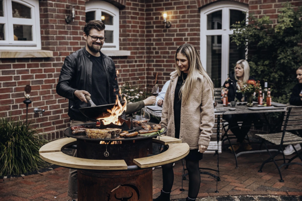 Mann grillt mit Flammen in Pfanne neben Frau im Garten bei geselligem Beisammensein