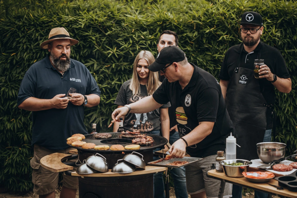 Gruppe von sechs Personen grillt gemeinsam im Freien, Männer und Frau bereiten Pizza und Fleisch zu