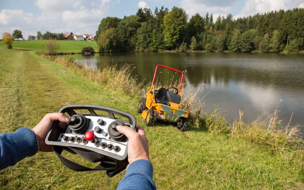 Person bedient ferngesteuerten Aufsitzmäher AS 940 Sherpa 4WD RC an einem sonnigen Tag neben einem Teich
