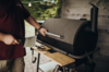 Mann bedient großen dunklen Grill im Garten mit Grillwerkzeugen auf Holztisch
