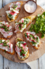 Bruschetta mit Frischkäse, Radieschen und Kräutern auf einem Holzbrett neben Meersalz und Feldsalat