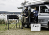 Zwei Personen entspannen neben einem Petromax Gastisch und einem Camping-Van am See