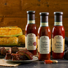 Drei Flaschen Stonewall Kitchen Barbecue-Saucen neben Grillgerichten auf Holztisch