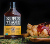 Flasche Rufus Teague Honey Sweet BBQ Sauce neben gegrilltem Hähnchenbein