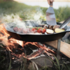 Person kocht Fleisch und Gemüse auf Petromax Pfannenknecht über Lagerfeuer