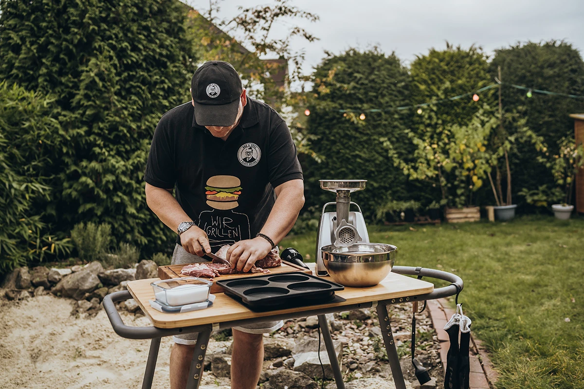 Mann bereitet Fleisch am Grilltisch im Garten vor, Grillwerkzeuge an Haken