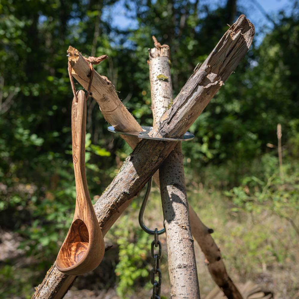 Petromax Kelle aus Olivenholz hängt an einem rustikalen Holzstativ im Wald