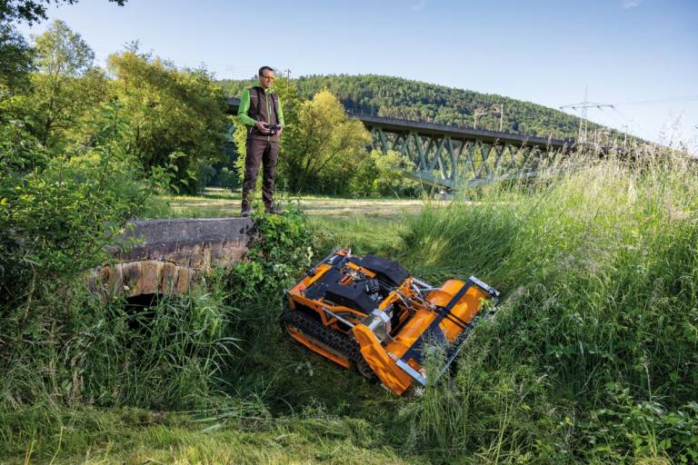 Mann bedient ferngesteuerten Schlegelmäher AS 1000 Ovis RC in grüner Landschaft unter Brücke
