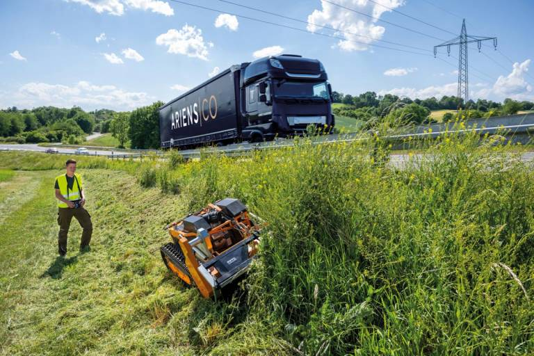 Mann bedient ferngesteuerten Schlegelmäher AS 1000 Ovis RC auf steiler Böschung neben Autobahn