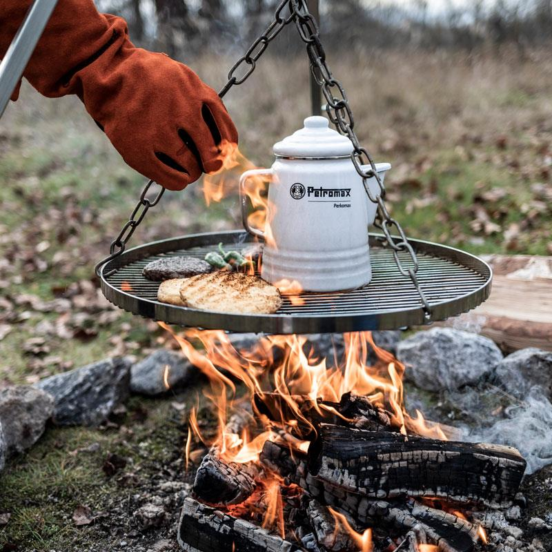Person verwendet Petromax Hänge-Rost über Lagerfeuer zum Grillen von Steak und Gemüse