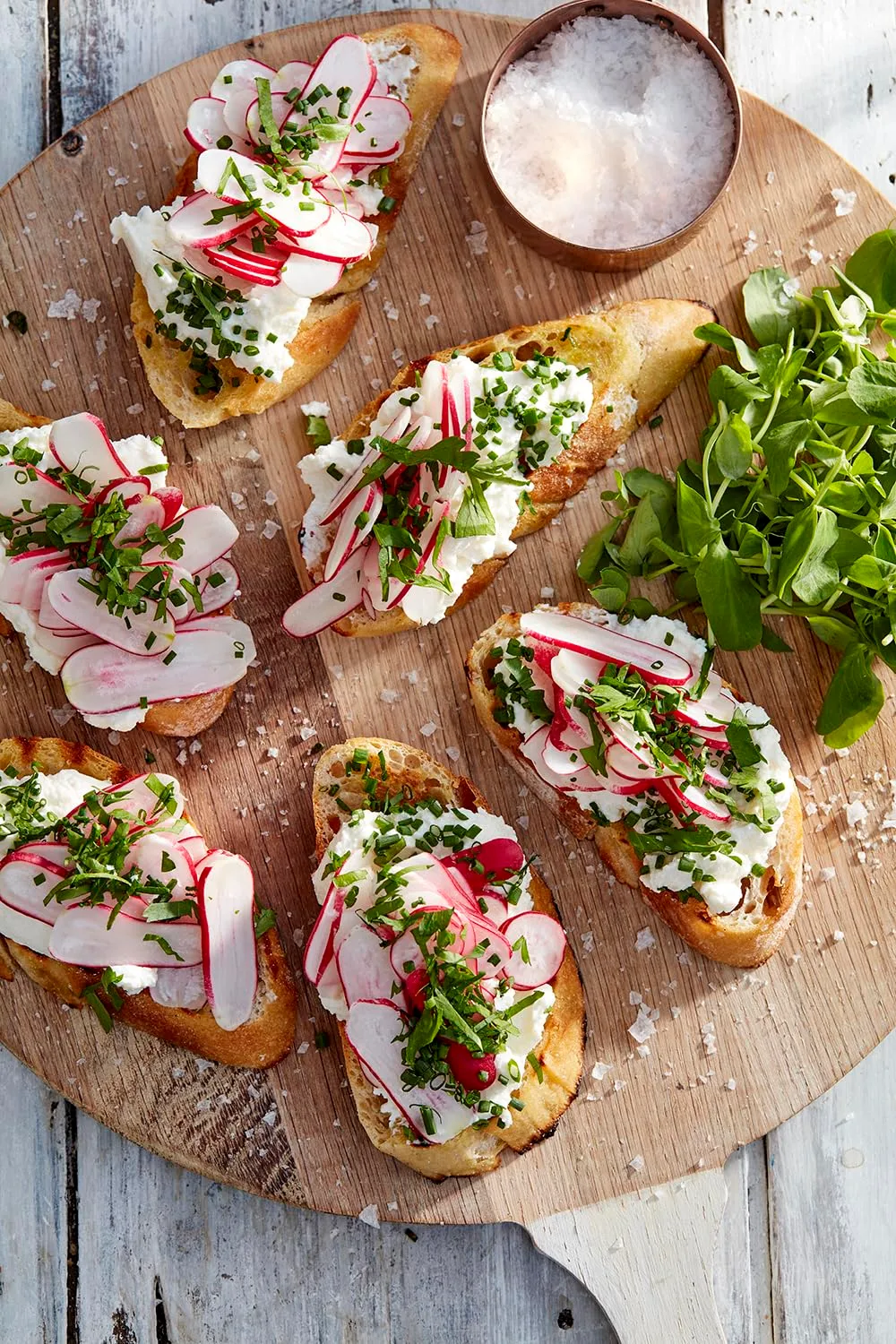 Bruschetta mit Frischkäse, Radieschen und Kräutern auf einem Holzbrett neben Meersalz und Feldsalat