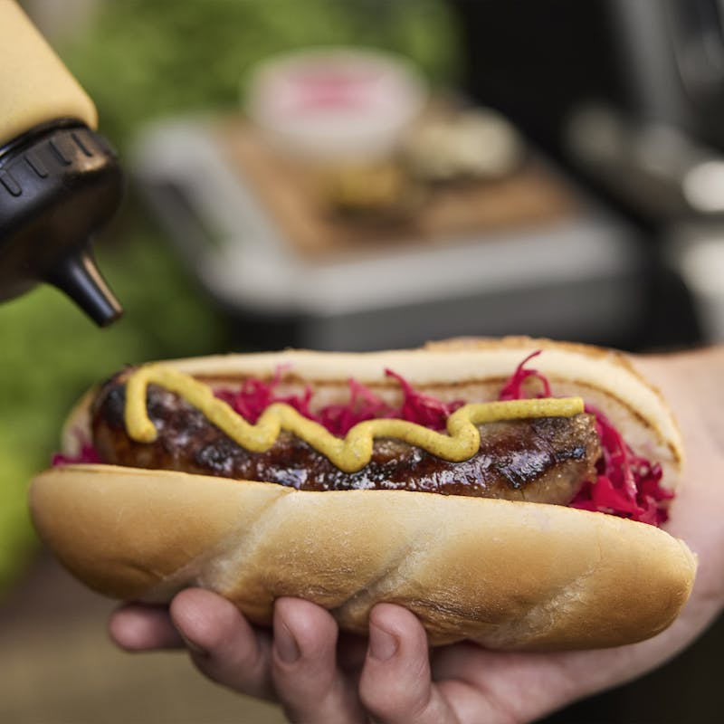 Hand hält appetitlichen Hotdog vor einem Gasgrill