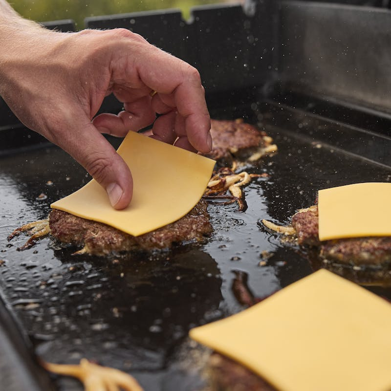Hand legt Käse auf Burger auf einer Grillplatte, Ölspritzer sichtbar