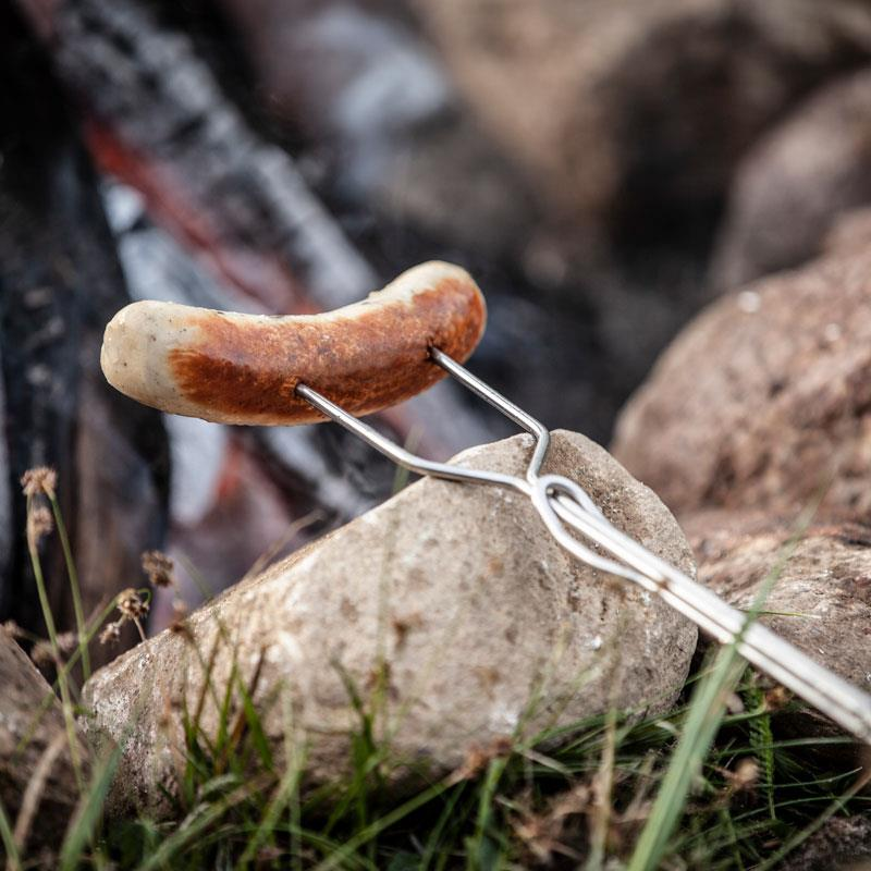 Petromax Lagerfeuer-Spieß ls1 mit Wurst über glühenden Kohlen im Freien