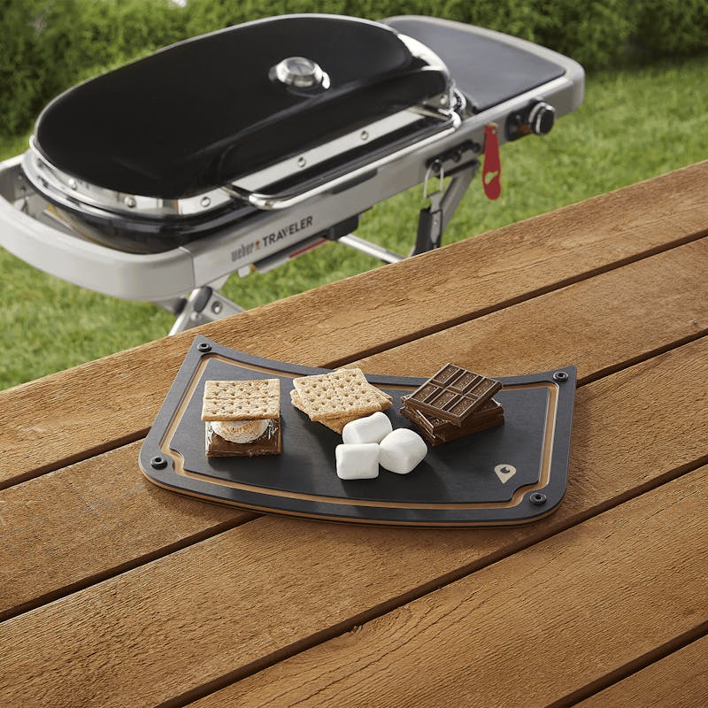 Weber Traveler Schneidebrett auf Holztisch mit S'mores Zutaten, Grill im Hintergrund
