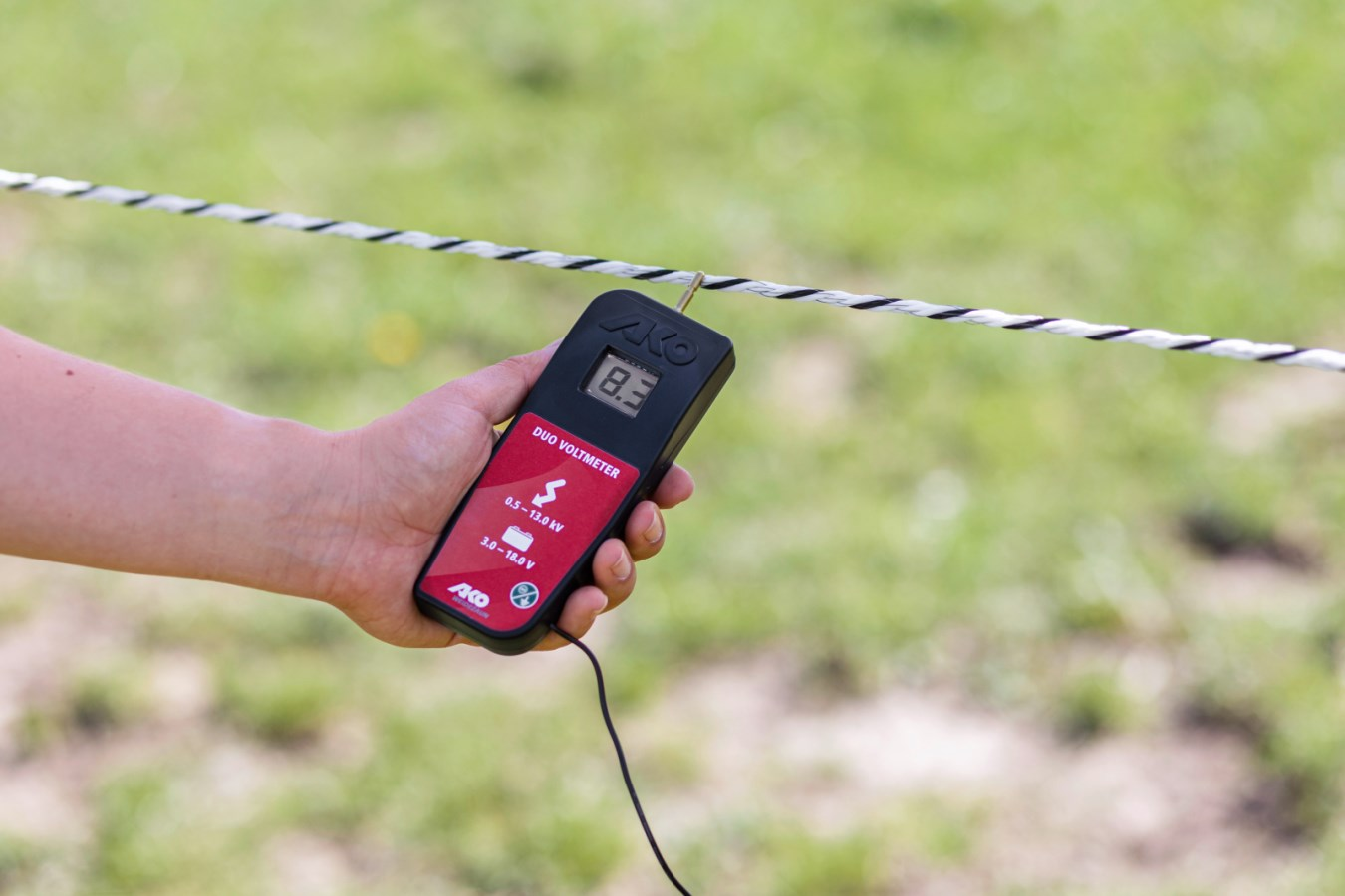 Hand hält AKO DUO-Voltmeter zur Messung der Spannung eines Elektrozauns auf grünem Hintergrund
