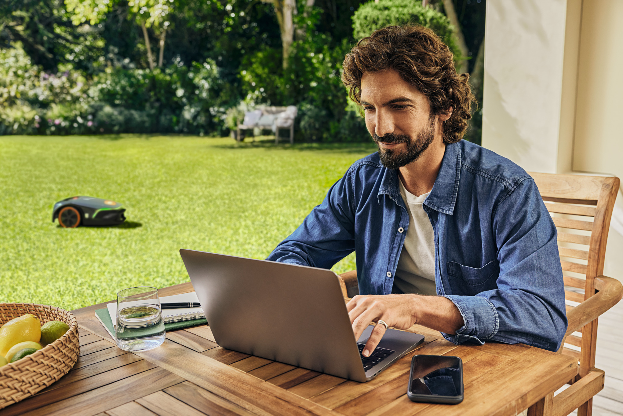 Mann arbeitet am Laptop im Garten mit STIHL Mähroboter im Hintergrund