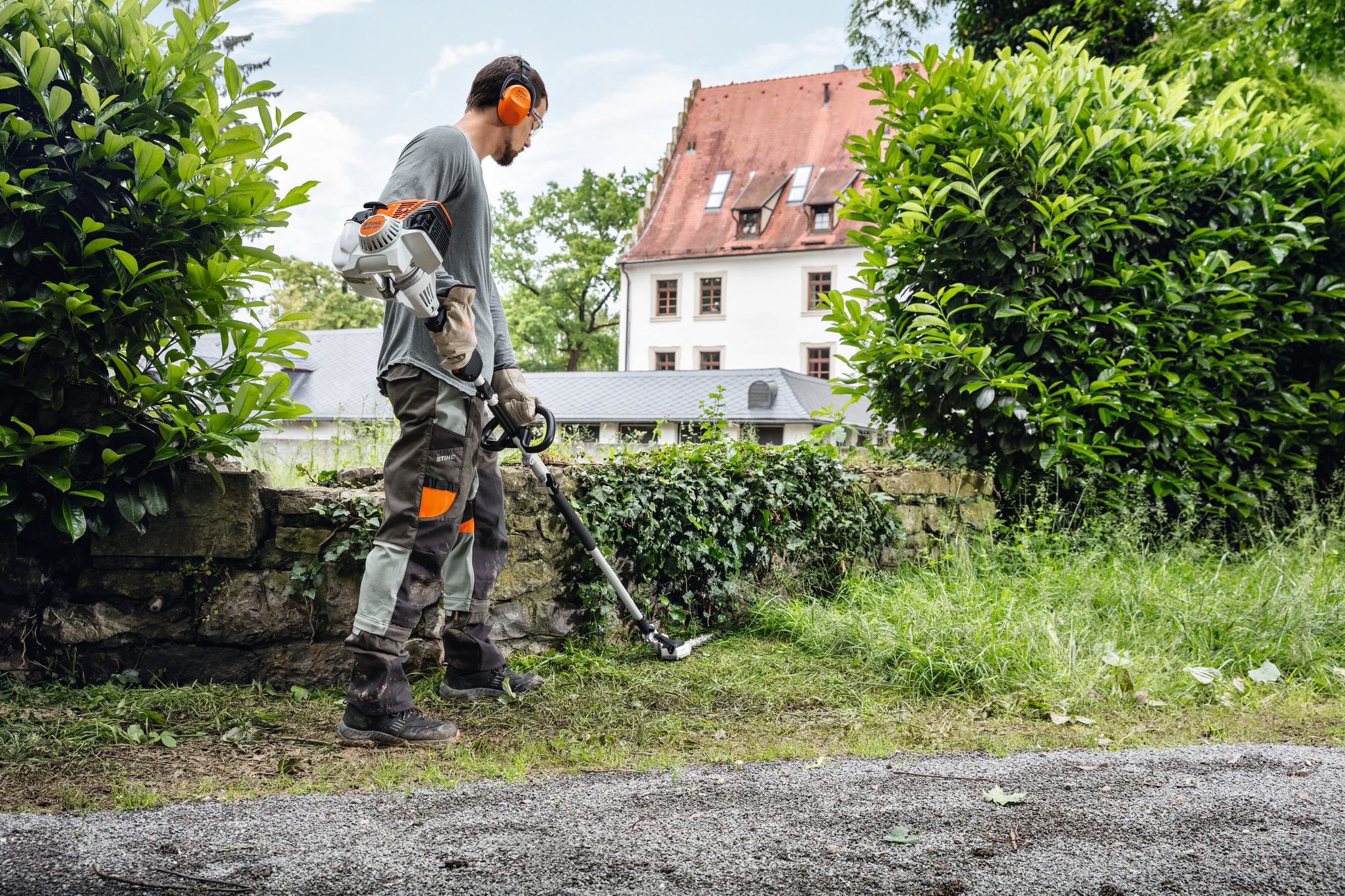 Mann verwendet Stihl Gestrüppschneider in Garten nahe Steinmauer und traditionellem Haus