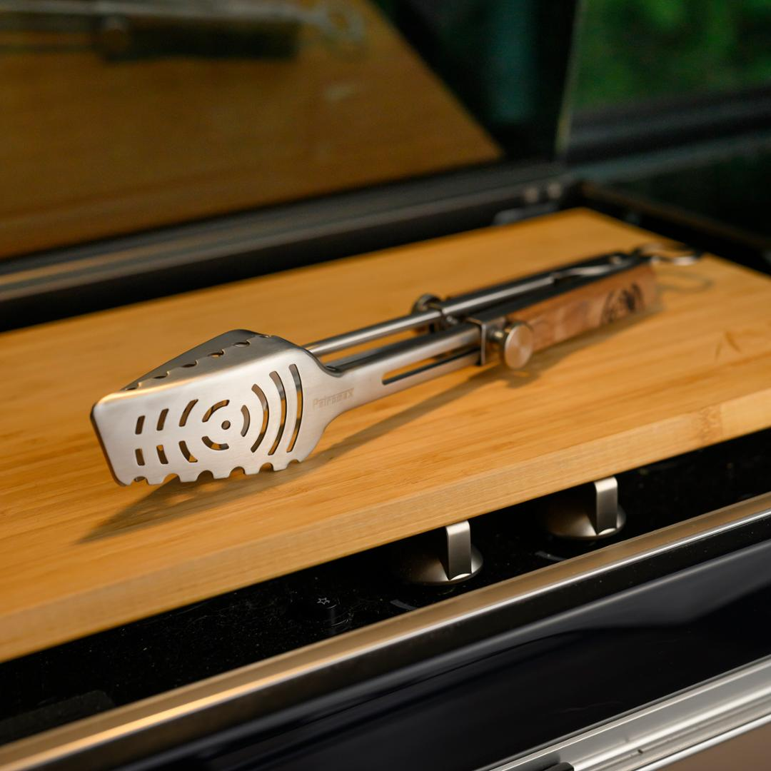 Teleskopische Grillzange auf Holzoberfläche neben einem Grill