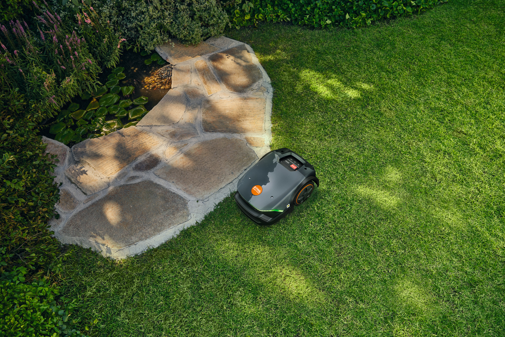 STIHL iMOW 3 EVO Mähroboter mäht Rasen neben steinernem Weg und Teich