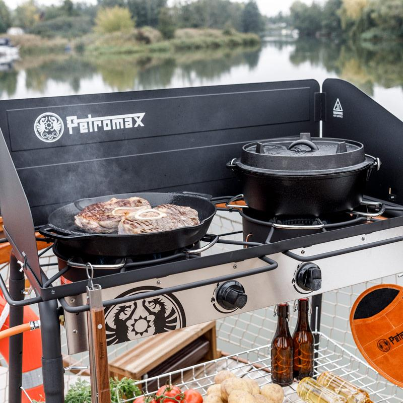 Petromax Gastisch mit Einzelbrenner im Einsatz im Freien, Steaks braten, Bier und Gemüse neben einem See