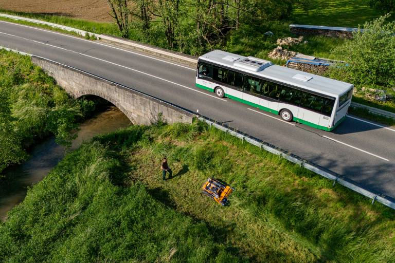 Bus auf Brücke über Fluss mit ferngesteuertem Mäher und Bediener auf Wiese