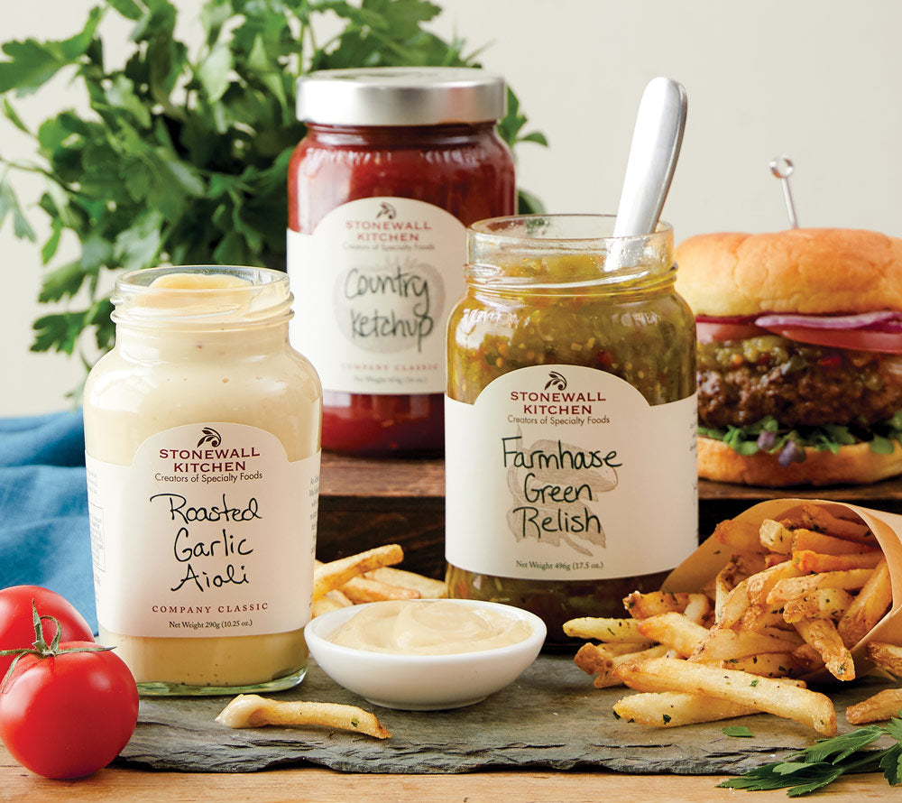 Stonewall Kitchen Saucen auf Schieferplatte mit Burger und Pommes im Hintergrund
